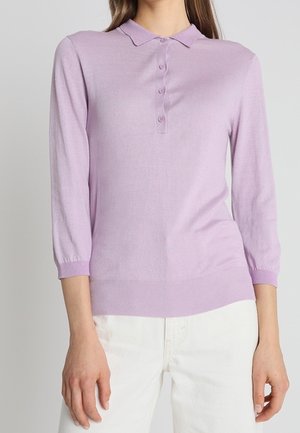 Kobieta w jasnofioletowej koszulce polo z długim rękawem i guzikami, w białych spodniach, stojąca na jednolitym tle.