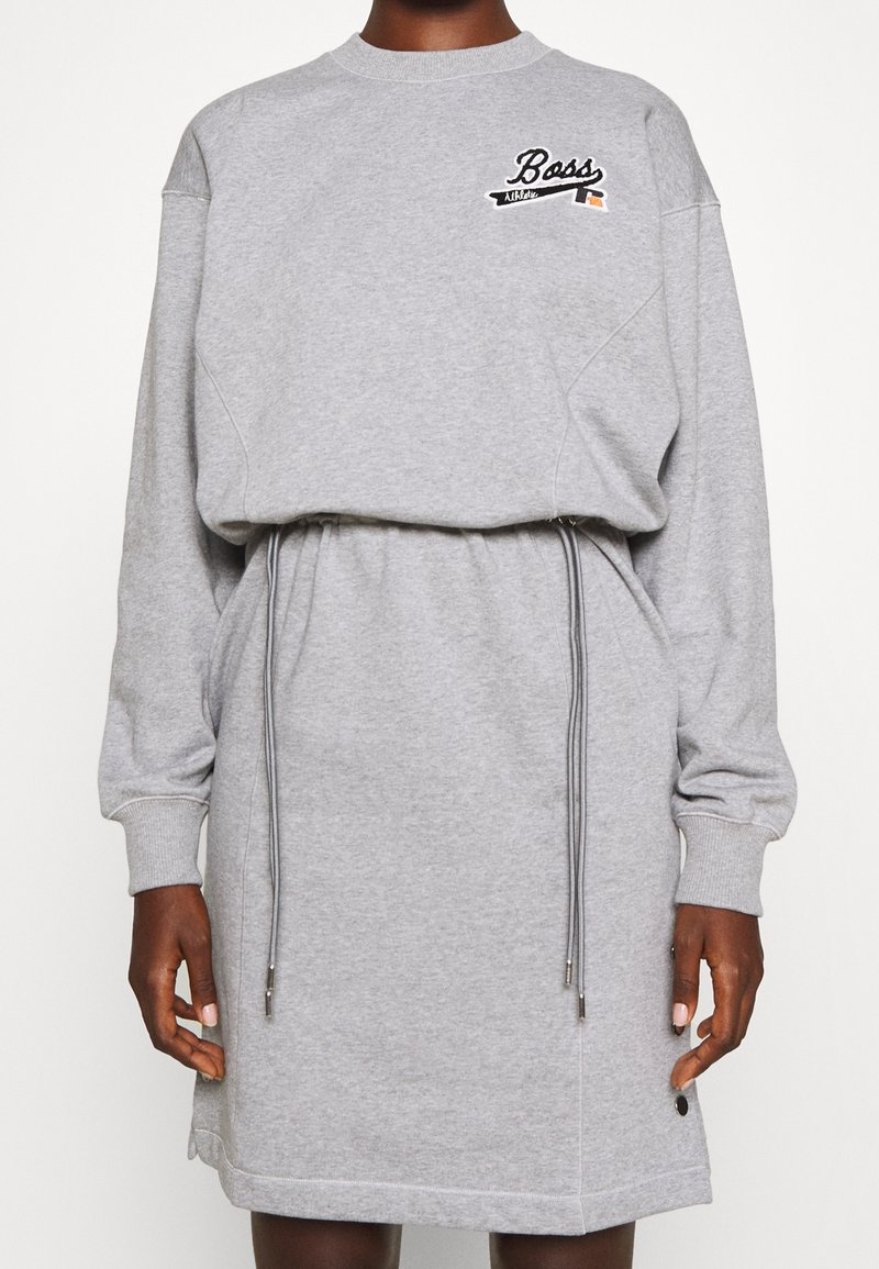 Robe sweat-shirt gris à manches longues avec taille à cordon et logo « Boss » sur la poitrine gauche, portée par une personne à la peau foncée.