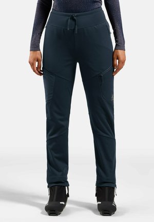 Person, die dunkelblaue, schlanke Outdoor-Hose mit Reißverschlusstaschen und schwarzen Winterstiefeln trägt, steht vor hellem Hintergrund.