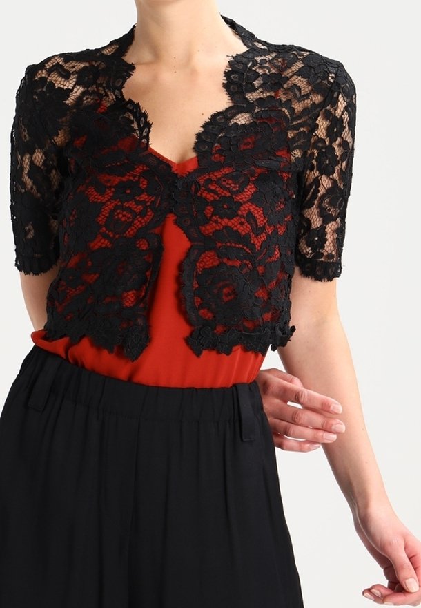 Femme portant un boléro court en dentelle noire transparente à manches courtes par-dessus un top rouge et un pantalon noir taille haute sur un fond uni.
