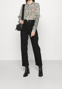 En kvinna står självklart, iklädd en blommig blus med volangärmar, svarta högmidjade jeans och klackstövlar, medan hon håller en svart väska.