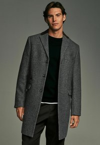 Grå frakke med fiskebensmønster og brune krave detaljer, der har tre knapper og klaplommer. Båret over en sort sweater.