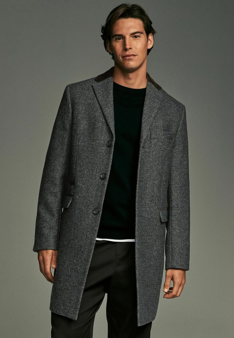 Grå frakke med fiskebensmønster og brune krave detaljer, der har tre knapper og klaplommer. Båret over en sort sweater.