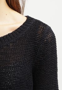 Pull noir en maille avec une texture ajourée, présentant un large col rond et des bordures côtelées au niveau du col et des manches.