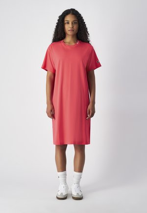 Koraal oversized t-shirt jurk, korte mouwen, ronde halslijn, zachte stof, logodetail nabij de schouder, gecombineerd met witte sokken en schoenen.