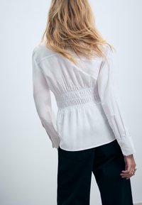 Donna con capelli biondi ondulati, che indossa una camicia bianca a maniche lunghe con vita arricciata e pantaloni neri, vista di spalle su uno sfondo semplice.
