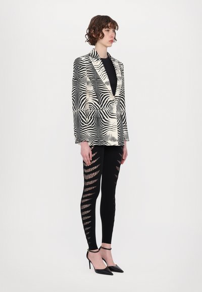 Zebrastribet blazer i sort og beige, parret med sorte leggings med udskæringsdetaljer. Modellen bærer sorte strophæle.