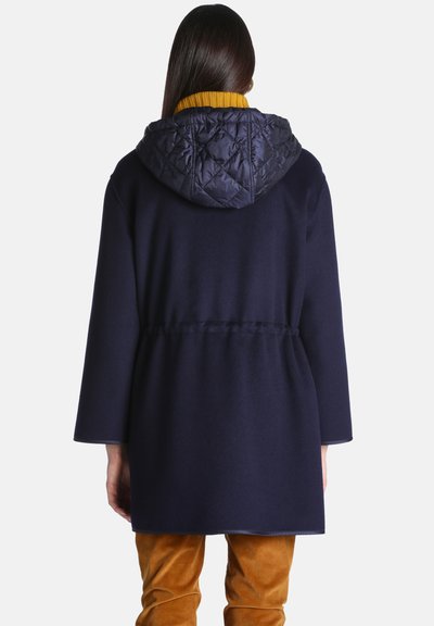 Femme vue de dos portant un manteau bleu marine avec une capuche matelassée, un pull à col roulé jaune moutarde et un pantalon marron.