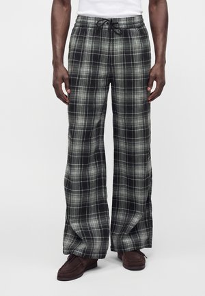 Homme portant un pantalon large à carreaux noir et blanc avec une ceinture élastique et un cordon noir, associé à des chaussures marron et un haut blanc.