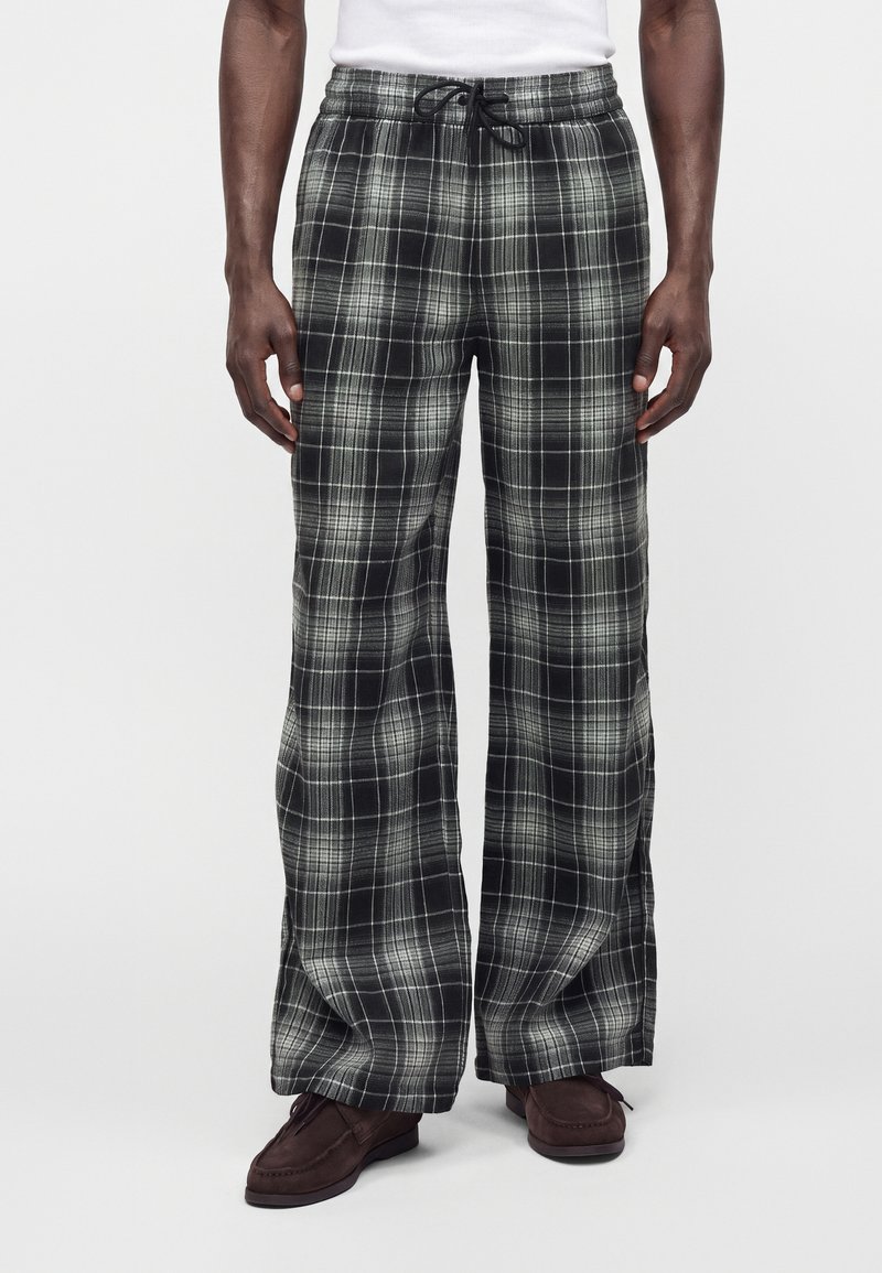 Homme portant un pantalon large à carreaux noir et blanc avec une ceinture élastique et un cordon noir, associé à des chaussures marron et un haut blanc.