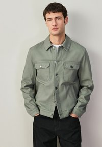 Jauns vīrietis ar īsiem tumšiem matiem, valkā gaiši olīvzaļu pogātu jaku pār baltiem krekls un melnām biksēm, rokas kabatās, neitrāla fona priekšā.