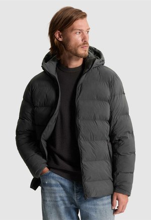 Homme portant une veste matelassée à capuche gris foncé par-dessus un pull noir et un jean bleu, regardant sur le côté avec une main dans la poche.