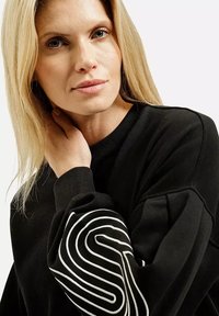 Czarny sweter z długimi rękawami, z charakterystycznym białym wzorem fali na lewym rękawie i ściągaczami. Gładka tekstura.