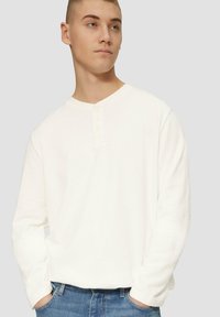 Pikkade varrukatega valge henley särk, mis on valmistatud tekstuurilisest kangast, ümarate krae ja neljast nööbist koosneva sisekeskkonna poolest. Koos siniste teksadega.