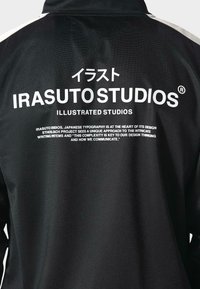 Czarna kurtka z białym tekstem i japońskimi znakami na plecach, podkreślająca "IRASUTO STUDIOS" oraz szczegóły dotyczące etosu projektowania.