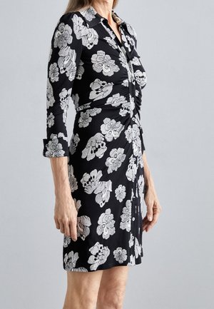 Vrouw in een zwarte jurk met witte bloemenpatronen, lange mouwen opgerold en een geplooide voorkant.