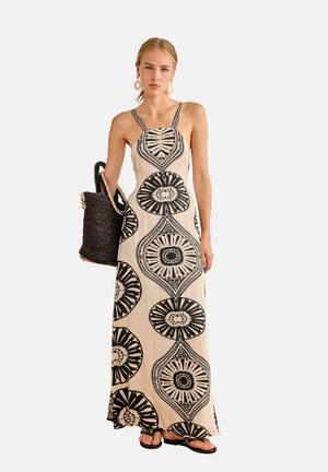 Mujer vestida con un vestido maxi sin mangas beige con grandes patrones circulares negros, sosteniendo un bolso tejido negro y con sandalias negras.