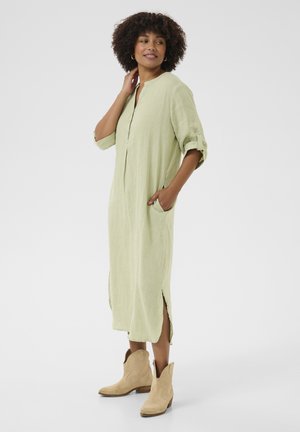 Robe en lin vert clair avec un col mandarin, des manches trois-quarts, des poches latérales et des fentes hautes des deux côtés, portée avec des bottes de cowboy beige.
