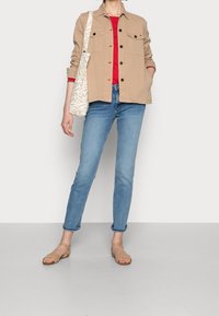 Beige kožená bunda přes červený top, světle modré džíny a béžové sandály. Obsahuje bílou tašku se světlem vzorem.