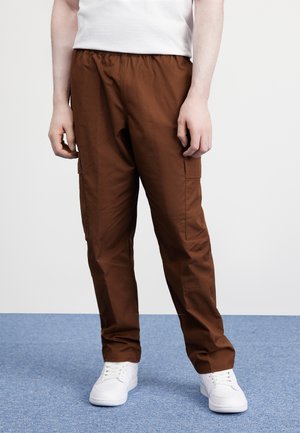 Personne vêtue d'un pantalon cargo marron et de baskets blanches, debout sur un tapis bleu, les mains détendues le long du corps.