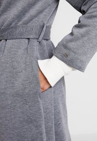 Šedý župan z měkké látky, který se zužuje v pase, s bočními kapsami a žebrovanými bílými manžetami s kovovým detailem u rukávu.