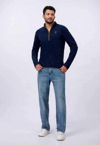 Navy fleece pullover with a zip neck, featuring a gold zipper and logo. Paired with light blue jeans and white sneakers.