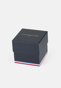 Boîte en carton noire avec un couvercle, présentant un logo blanc "TOMMY HILFIGER" au-dessus de "WATCHES." Accents rouge, blanc et bleu à la base.