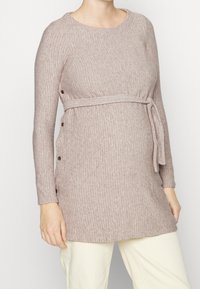 Schwangere Frau trägt einen gerippten, langärmeligen beigen Strick-Maternity-Pullover mit seitlichen Knöpfen und einem gebundenen Gürtel über weißen Hosen.