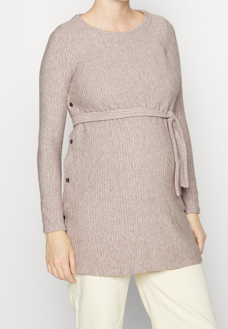 Schwangere Frau trägt einen gerippten, langärmeligen beigen Strick-Maternity-Pullover mit seitlichen Knöpfen und einem gebundenen Gürtel über weißen Hosen.