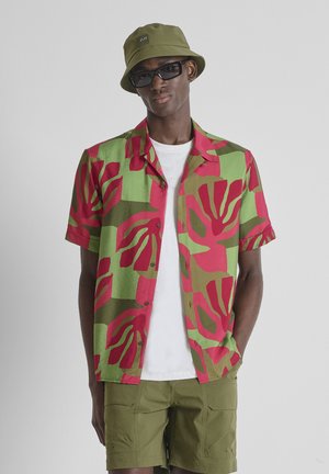 Hombre con camisa de manga corta con botones y estampado en verde y rojo, camiseta blanca, pantalones cortos verde oliva, sombrero tipo bucket verde oliva y gafas de sol negras.