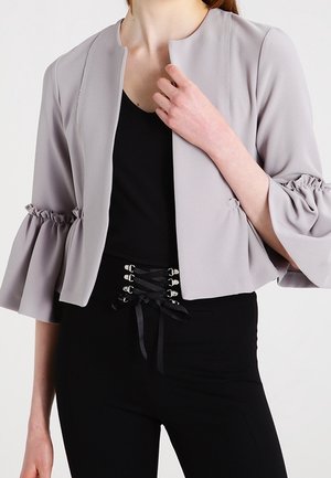 Nainen pukeutuneena vaaleanharmaaseen avoimeen takkiin, jossa on rypyttäytyneet 3/4-hihat, mustan topin ja korkeavyötäröisten mustien pitsikoristeisten housujen kanssa.