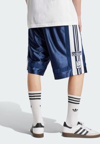 Calções de basquetebol em satin azul-marinho com riscas brancas laterais e o logo da Adidas, combinados com meias brancas com riscas pretas e ténis com detalhes pretos.