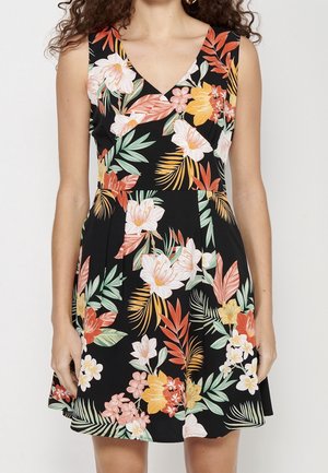 Czarna sukienka bez rękawów z głębokim dekoltem w kształcie litery V i kolorowym tropikalnym kwiatowym wzorem w odcieniach pomarańczowego, żółtego, białego i zielonego, noszona przez stojącą osobę.