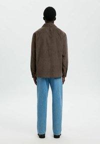 Persona de pie de espaldas, vistiendo una camisa de pana marrón, jeans azul claro y zapatos negros, frente a un fondo claro y liso.