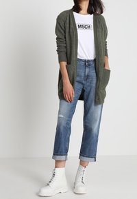 Cardigan lungo verde oliva con maniche a coste, t-shirt bianca con grafica, jeans chiari strappati e stivaletti bianchi con lacci.