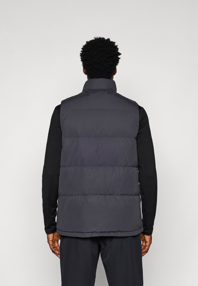 Graue Pufferweste mit hohem Kragen, die genähte Abschnitte und eine elegante, glatte Textur aufweist. Über einem taillierten schwarzen Langarmshirt getragen.