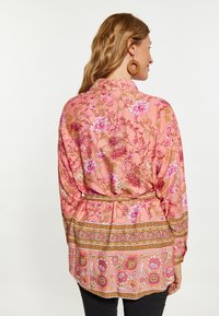 Blusa floreale rosa con maniche lunghe, caratterizzata da un orlo con motivo dorato e un nastro in vita. Tessuto morbido con dettagli floreali intricati.