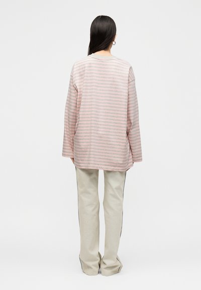 Άτομο με μακριά σκούρα μαλλιά που φοράει ένα άνετο ροζ και γκρι ριγέ μακρυμάνικο τοπ και μπεζ παντελόνια, στέκεται πλάτη σε λευκό φόντο.