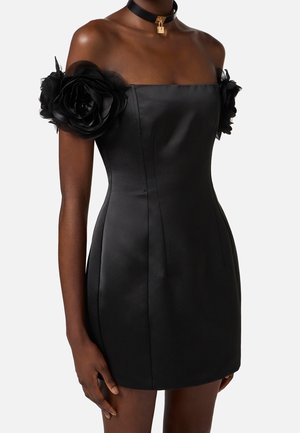 Kobieta ubrana w czarną satynową mini sukienkę z odkrytymi ramionami, dużymi kwiatowymi rękawami oraz czarny naszyjnik typu choker z złotą zawieszką w kształcie zamka.