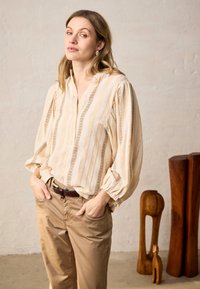 Femme aux cheveux ondulés portant une blouse beige à rayures et un pantalon marron clair, debout les mains dans les poches près de sculptures en bois posées au sol.