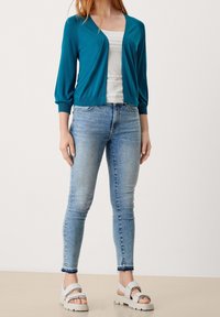 Tyrkysový cardigan s výstřihem do V a tříčtvrtečními rukávy, nošený přes bílé tričko, doplněný světle modrými skinny džíny a robustními sandály.