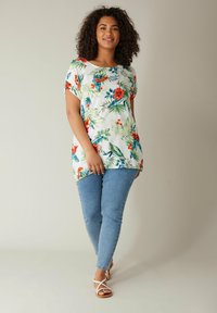 Florale Tunika aus weißem Stoff mit lebhaften roten und blauen Blumen, kombiniert mit hellblauen Skinny Jeans und weißen Riemchensandalen.