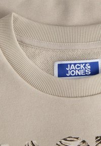Felpa di cotone beige con collo tondo a coste, con etichetta "JACK & JONES" in blu. Design semplice con texture liscia e motivo minimale.