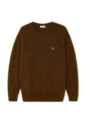 Un homme se tient face à nous, portant un pull marron, un pantalon en velours côtelé bordeaux et des baskets blanches sur un fond clair uni.