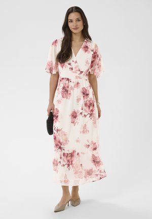 Femme en robe midi blanche à motif floral rose, manches flottantes, tenant une pochette noire tissée, portant des talons pointus beige.