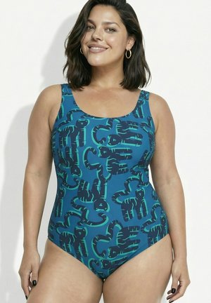 Donna con capelli castani alla lunghezza delle spalle che indossa un costume intero blu con forme astratte di animali verde scuro, sorridente verso la fotocamera.