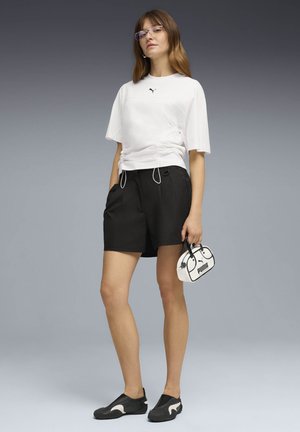 Mladá žena v bielom tričku Puma, čiernych šortkách, čiernych slip-on topánkach, s priehľadnými okuliarmi, držiaca malú bielu kabelku Puma na sivom pozadí.