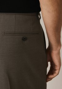 Braune maßgeschneiderte Hosen mit strukturiertem Finish, ausgestattet mit einer Gesäßtasche mit einem einzelnen schwarzen Knopf und einem schmalen Schnitt.