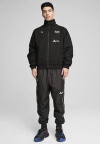 En ung mand står iført en sort Puma BMW Motorsport jakke og bukser med sorte sneakers foran en ensfarvet grå baggrund.