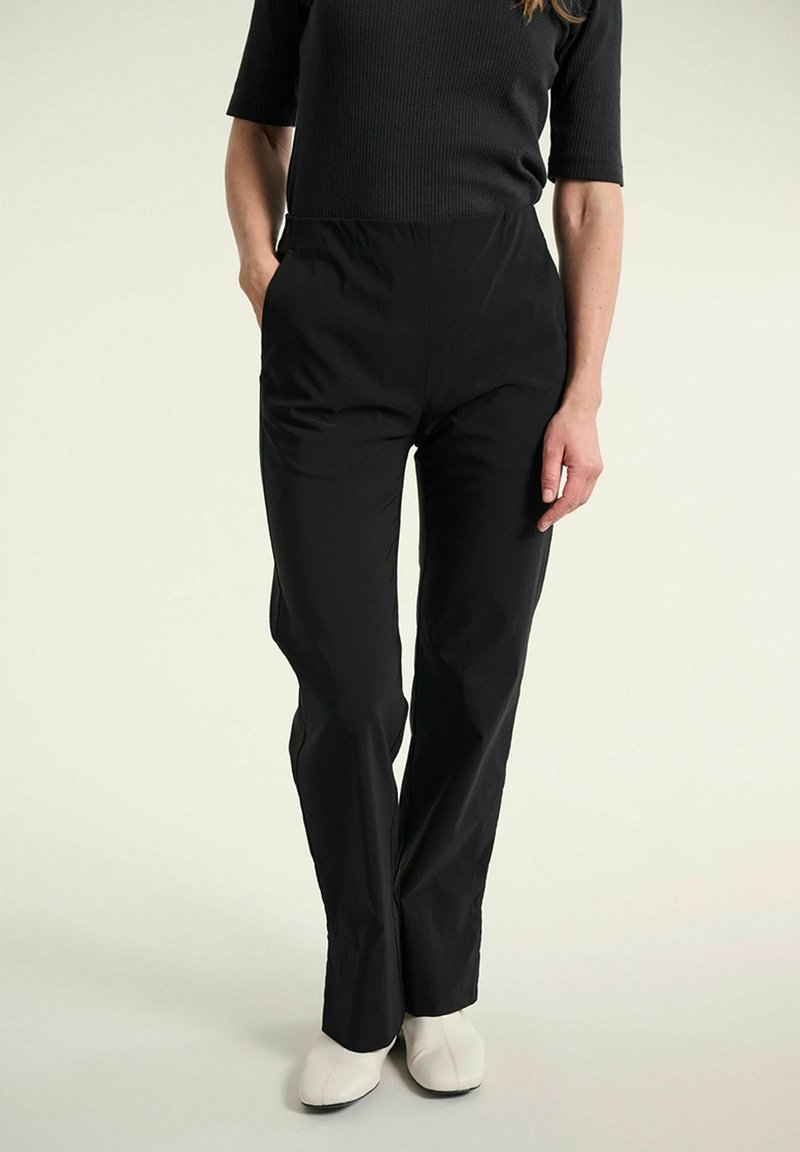 Femme portant un pantalon noir taille haute à jambes droites, mains dans les poches, et des bottines blanches à talons sur un fond uni.